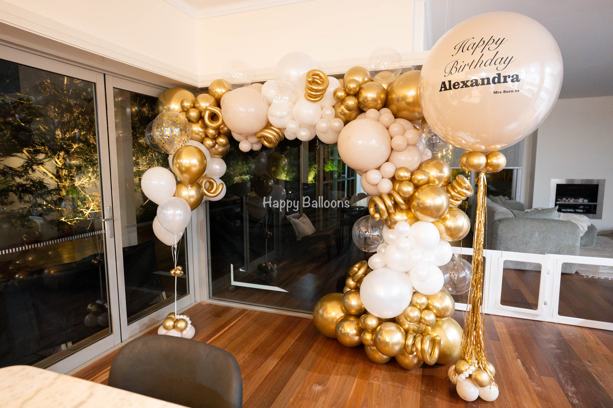 Wide view of a gold, cream and white organic balloon garland framing glass doors, with a personalised statement balloon and helium balloons.