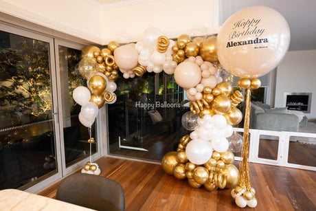 Wide view of a gold, cream and white organic balloon garland framing glass doors, with a personalised statement balloon and helium balloons.