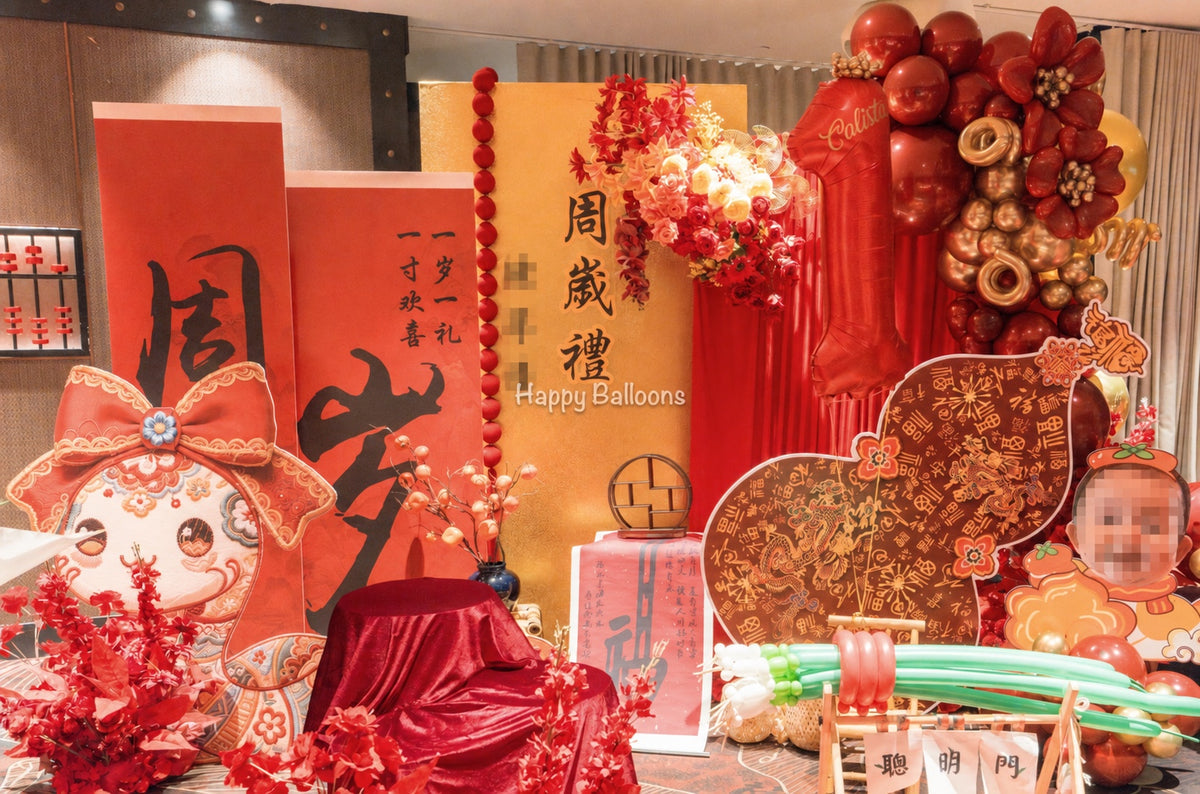 Wide view of a traditional Chinese first-birthday decoration setup with red and gold balloons, a draped backdrop, Chinese floral arrangement, child-inspired artwork and a 抓周 display.